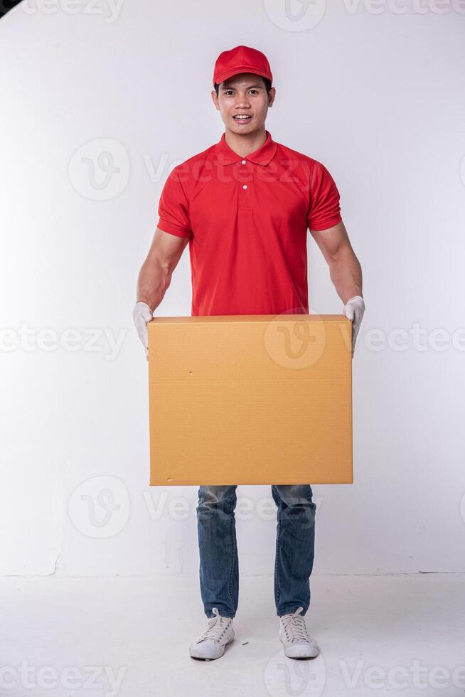 Immagine di un' contento giovane consegna uomo nel rosso berretto vuoto maglietta uniforme in piedi con vuoto Marrone cartone scatola isolato su leggero grigio sfondo studio foto