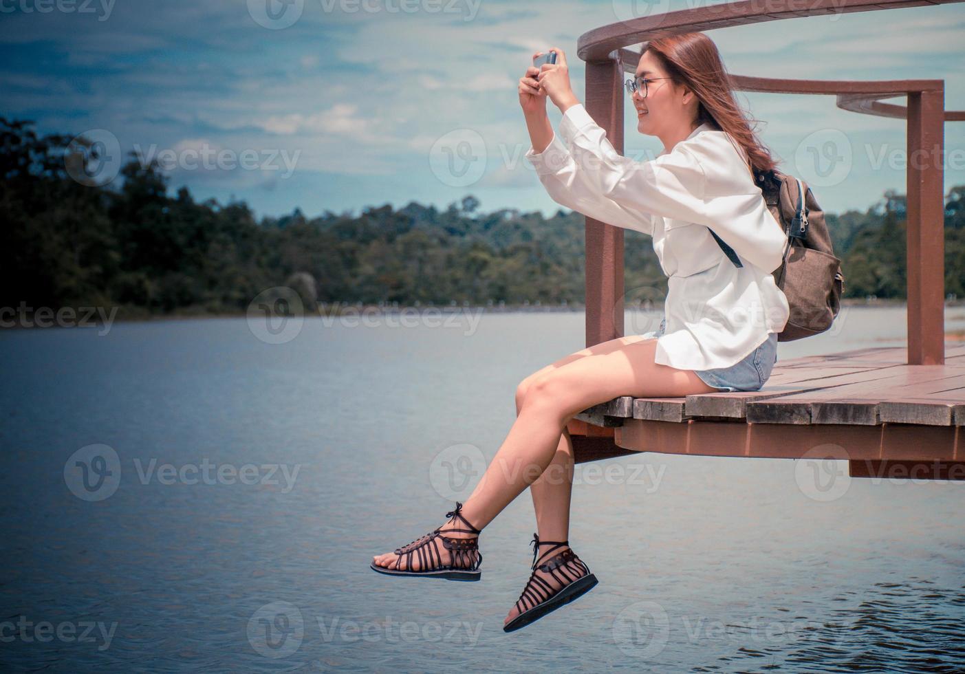 bellissimo asiatico donna indossare bicchieri e zaino per viaggio il vacanze. lei era indossare un' bianca camicia seduta su il ponte. utilizzando mobile Telefono per prendere immagini di fiume e montagne sentire rilassato. foto
