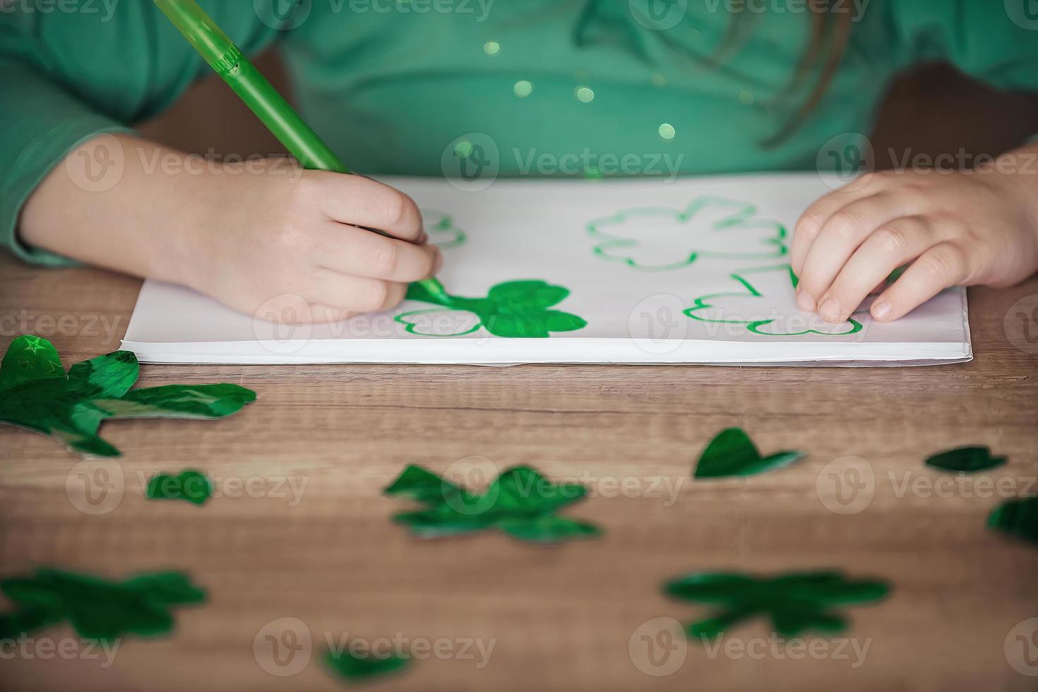 un' poco ragazza con un' bendare su sua testa disegna e tagli verde quadrifogli per st. Patrick giorno a un' tavolo a casa nel il cucina, Il prossimo per sua è sua bellissimo gatto con un' verde arco cravatta in giro il suo collo foto