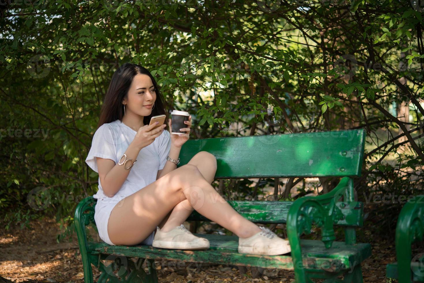 ritratto di una donna casual felice seduta su una panchina con caffè e telefono nel parco foto