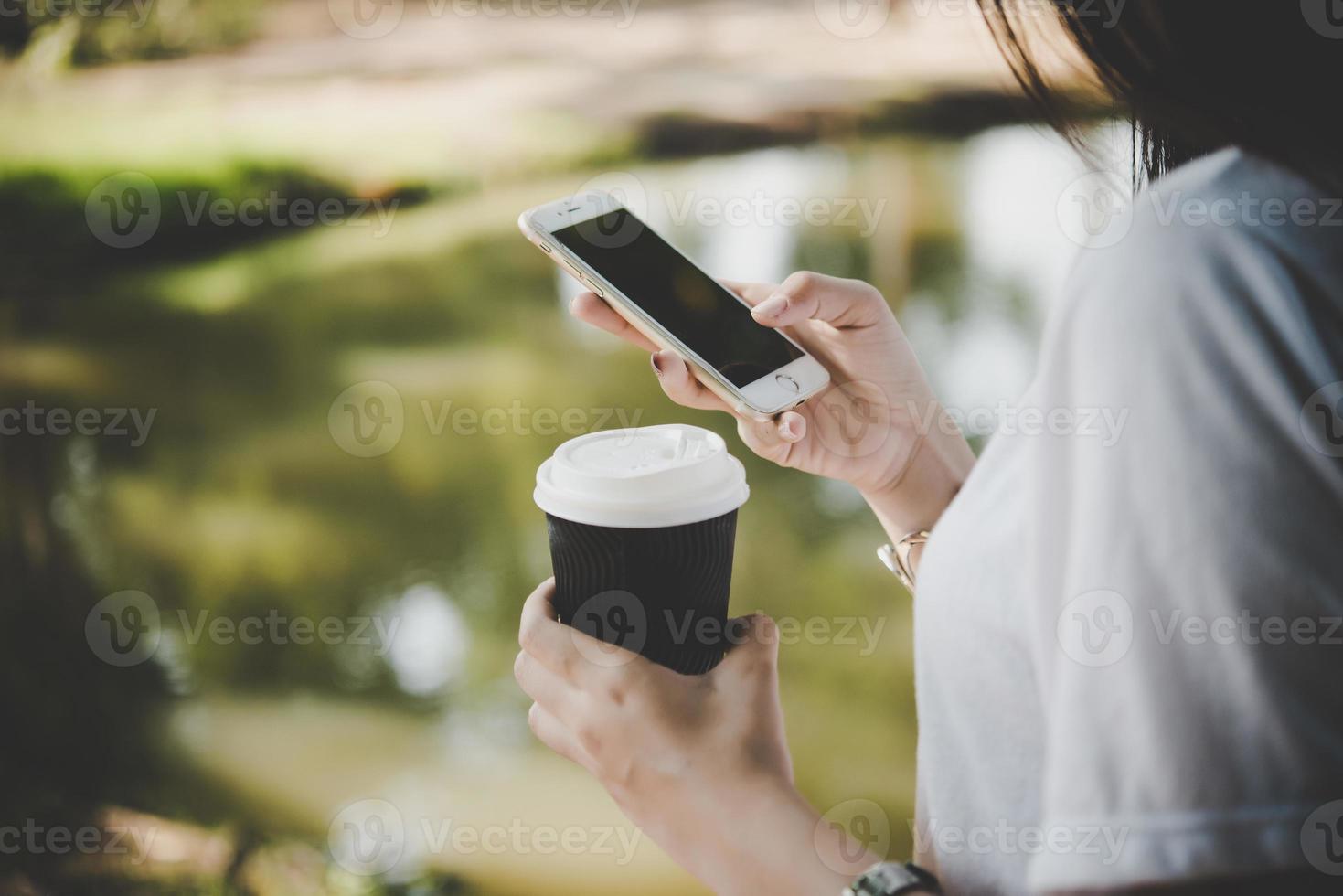 giovane donna che tiene tazza di caffè usa e getta durante la messaggistica di testo tramite smartphone all'aperto foto