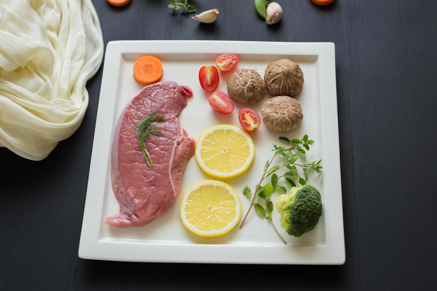 bistecca cruda, prezzemolo, funghi aneto, carote e pomodori a fette sul piatto bianco sul tavolo di legno nero foto