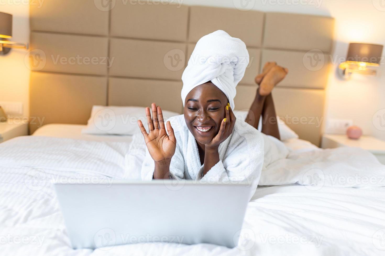 giovane nero donna nel asciugamano e accappatoio seduta su letto a casa.  bellissimo africano giovane donna indossare un' bianca accappatoio, con un' asciugamano  su sua testa nel letto a casa 20419002 Stock