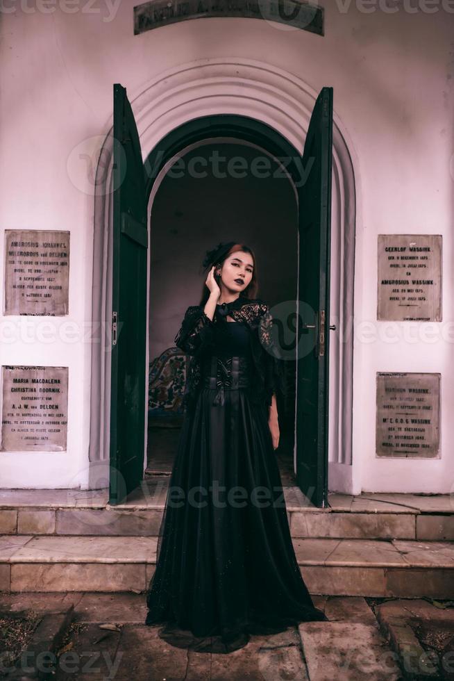 un' strega nel un tutti nero vestito e pauroso trucco era in piedi a il cimitero cancello foto