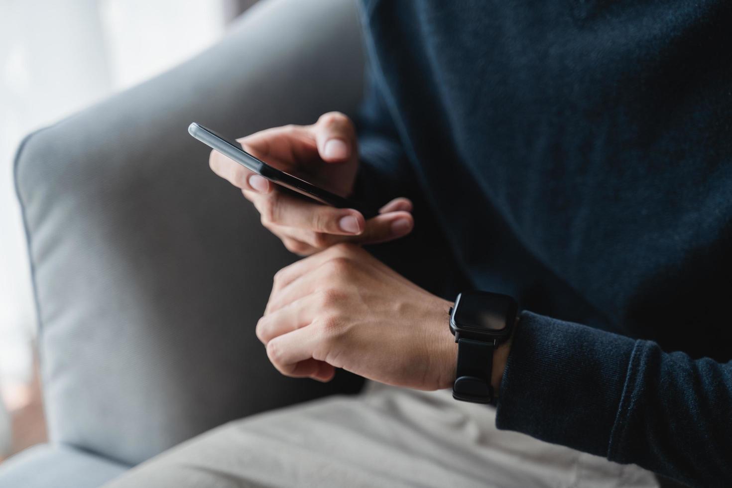 uomo utilizzando smartphone e orologio intelligente per puntamento attività su divano nel vivente camera a casa, senza fili connessione fra il orologio e mobile Telefono. foto