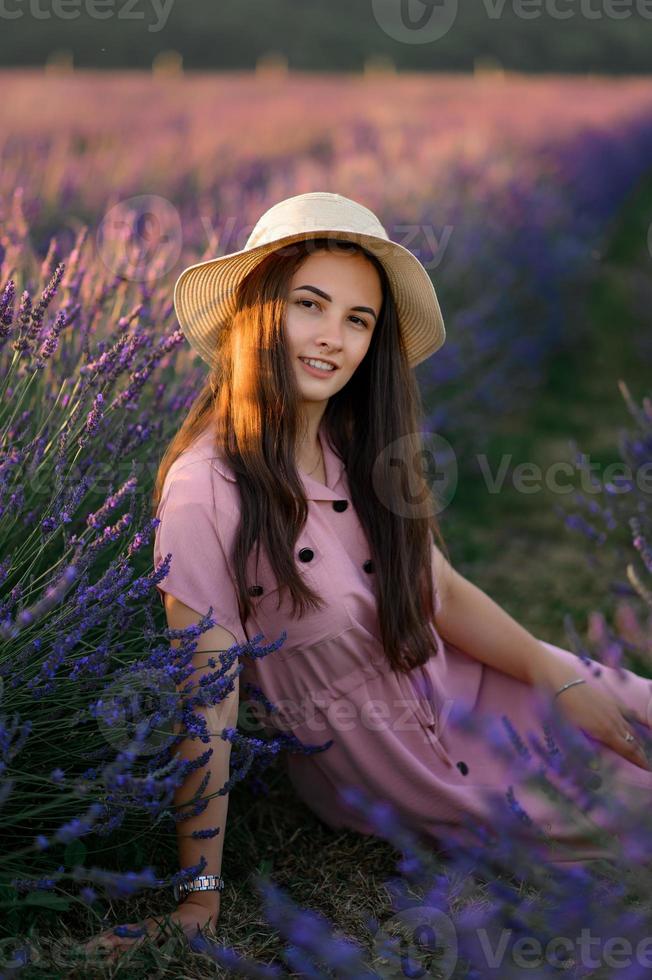 un' allegro giovane ragazza nel un' rosa vestito e un' cannuccia cappello nel sua mani sta tra lavanda cespugli. tramonto. foto