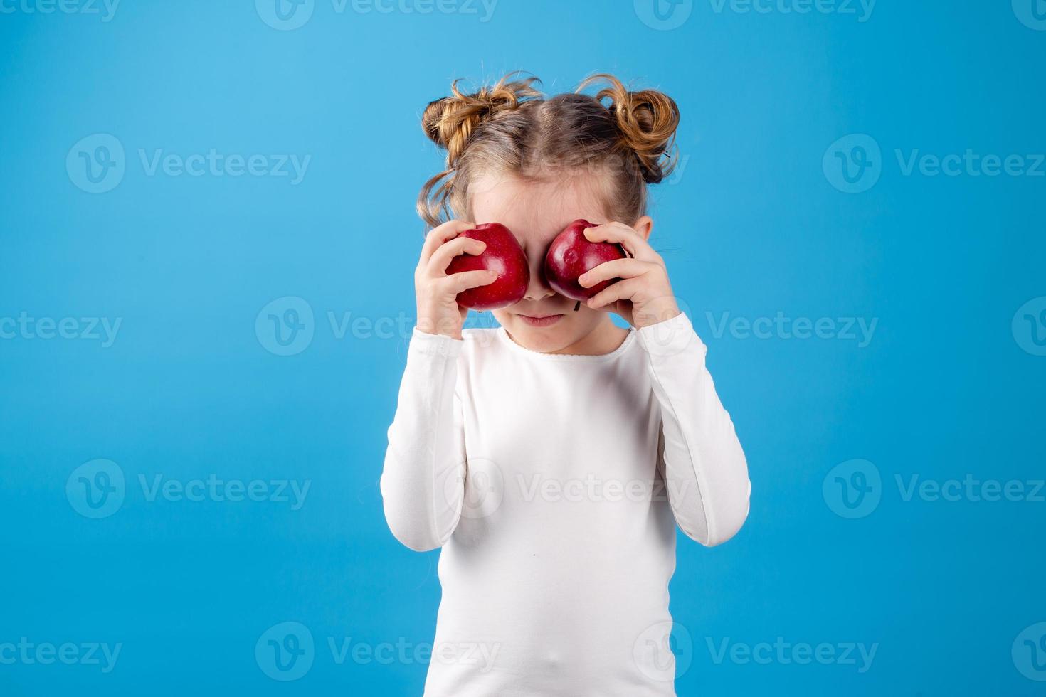 carino poco ragazza con riccioli nel un' a strisce vestito è Tenere un' grande rosso Mela nel sua mani. blu sfondo. utile prodotti per bambini. salutare merenda. spazio per testo. alto qualità foto