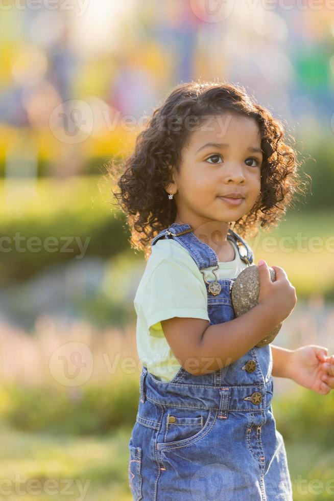 ritratto poco contento giocoso bambino piccolo bruno ragazza nel un' denim prendisole in piedi nel il giardino su un' soleggiato giorno. a piedi nel il fresco aria. concetto di un' contento infanzia. spazio per testo. alto qualità foto