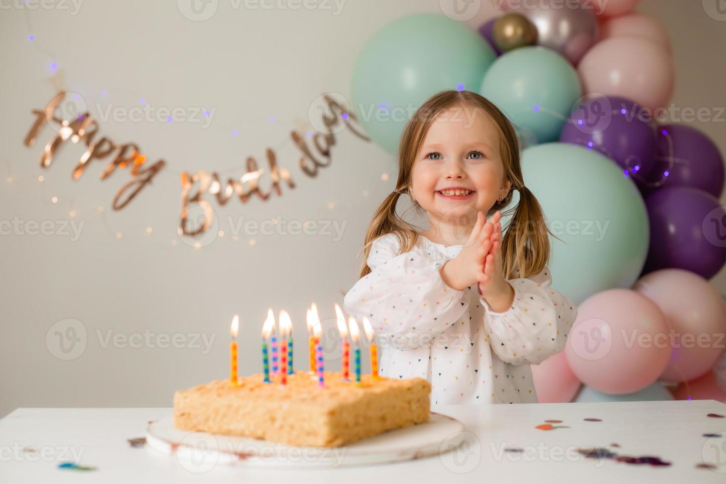 carino poco ragazza colpi su candele su un' compleanno torta a casa contro un' fondale di palloncini. del bambino compleanno foto