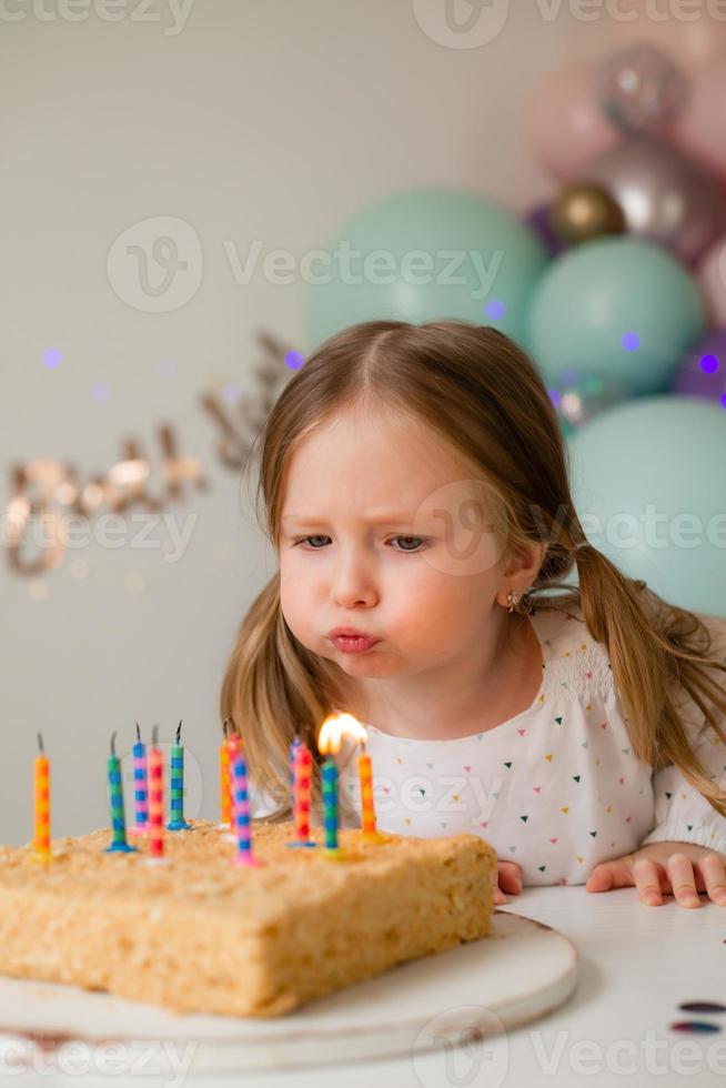 carino poco ragazza colpi su candele su un' compleanno torta a casa contro un' fondale di palloncini. del bambino compleanno foto