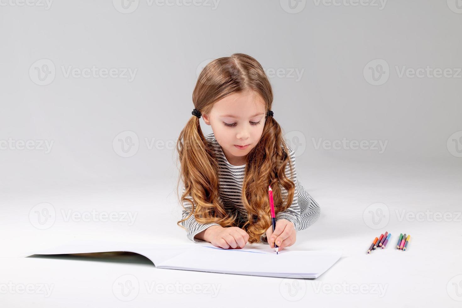 poco ragazza nel un' a strisce vestito disegna nel un album dire bugie su il pavimento. bambino sviluppo. stile di vita. prescolastico e scuola formazione scolastica. spazio per testo. alto qualità foto