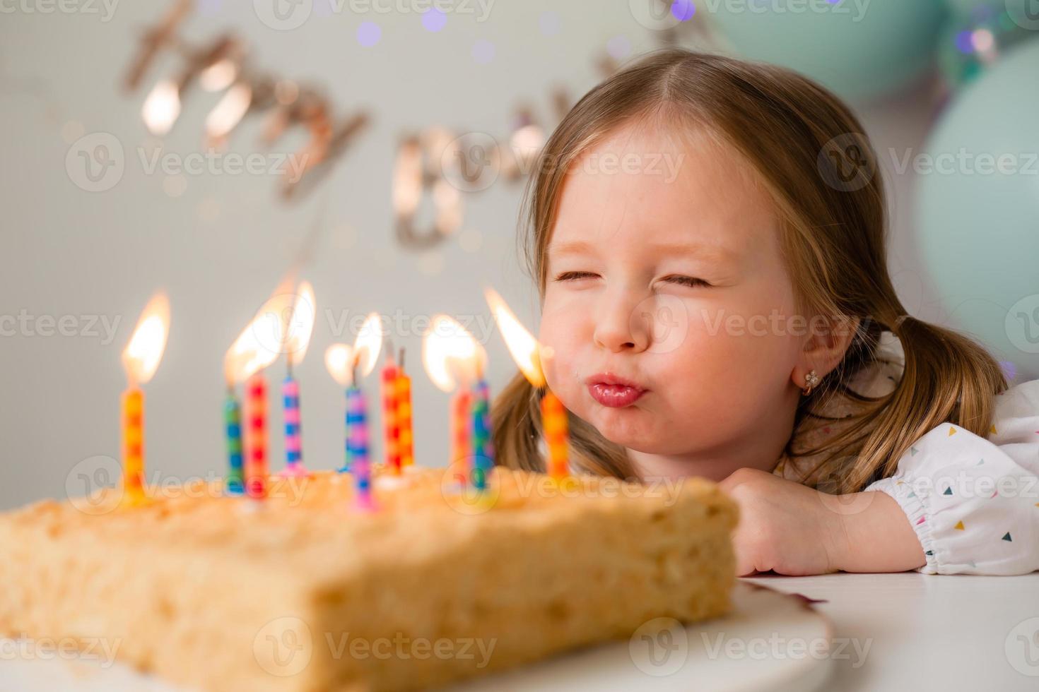 carino poco ragazza colpi su candele su un' compleanno torta a casa contro un' fondale di palloncini. del bambino compleanno foto