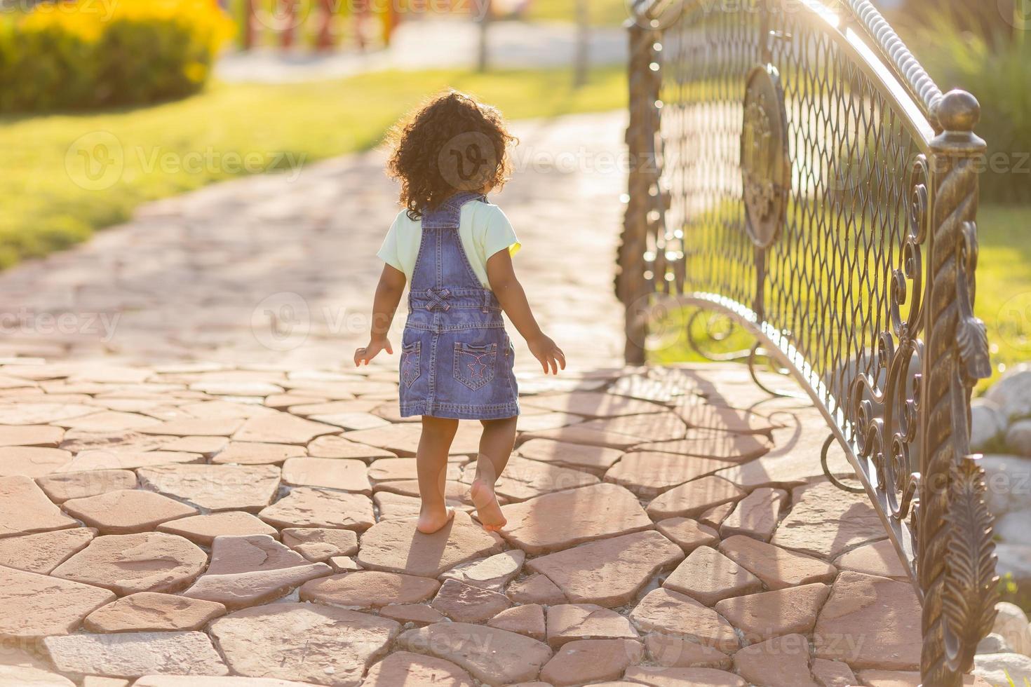 ritratto poco contento giocoso bambino piccolo bruno ragazza nel un' denim prendisole in piedi nel il giardino su un' soleggiato giorno. a piedi nel il fresco aria. concetto di un' contento infanzia. spazio per testo. alto qualità foto