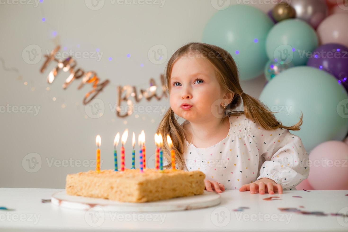 carino poco ragazza colpi su candele su un' compleanno torta a casa contro un' fondale di palloncini. del bambino compleanno foto