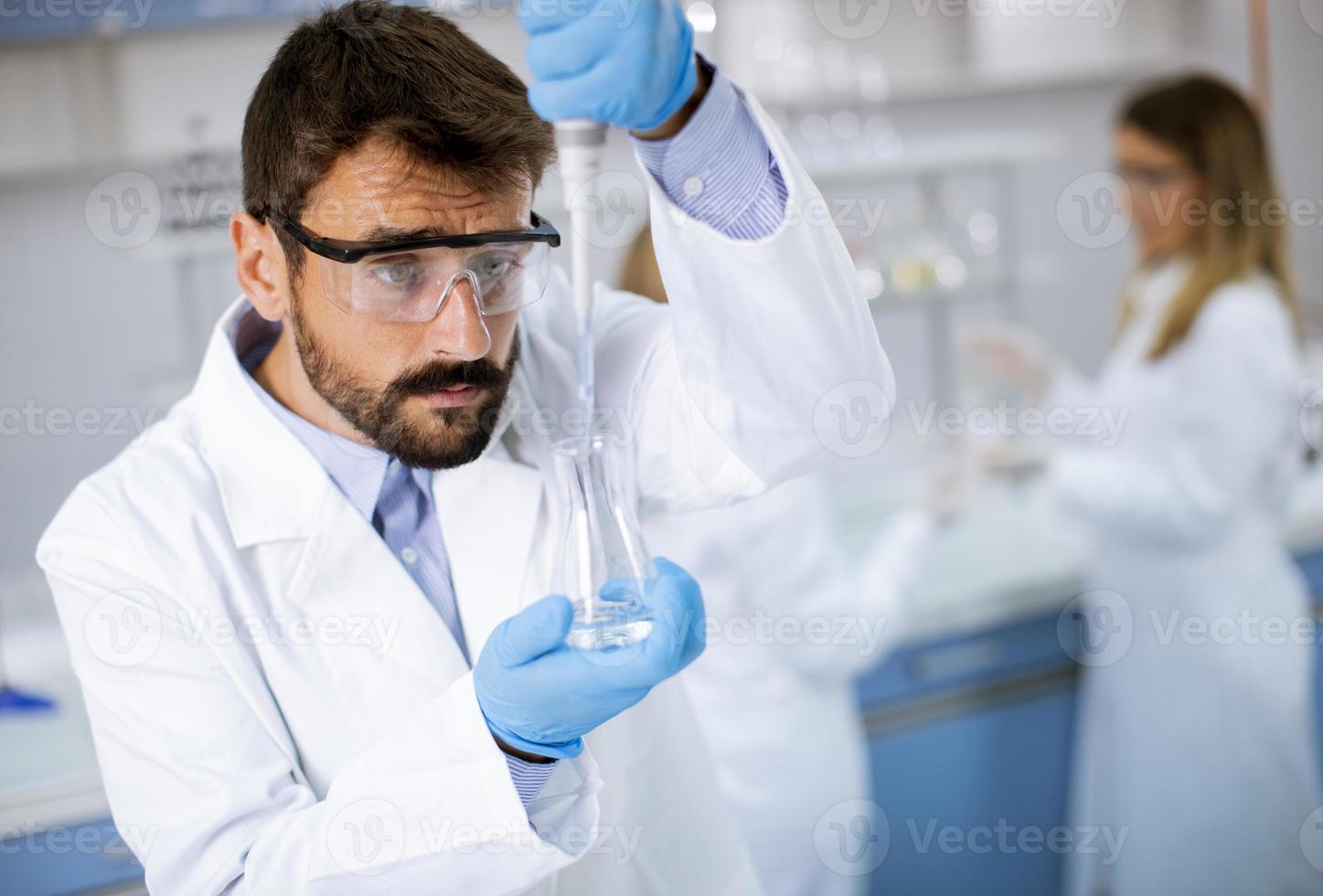 giovane ricercatore in indumenti da lavoro protettivi in piedi in laboratorio e analizzando il pallone con liquido foto