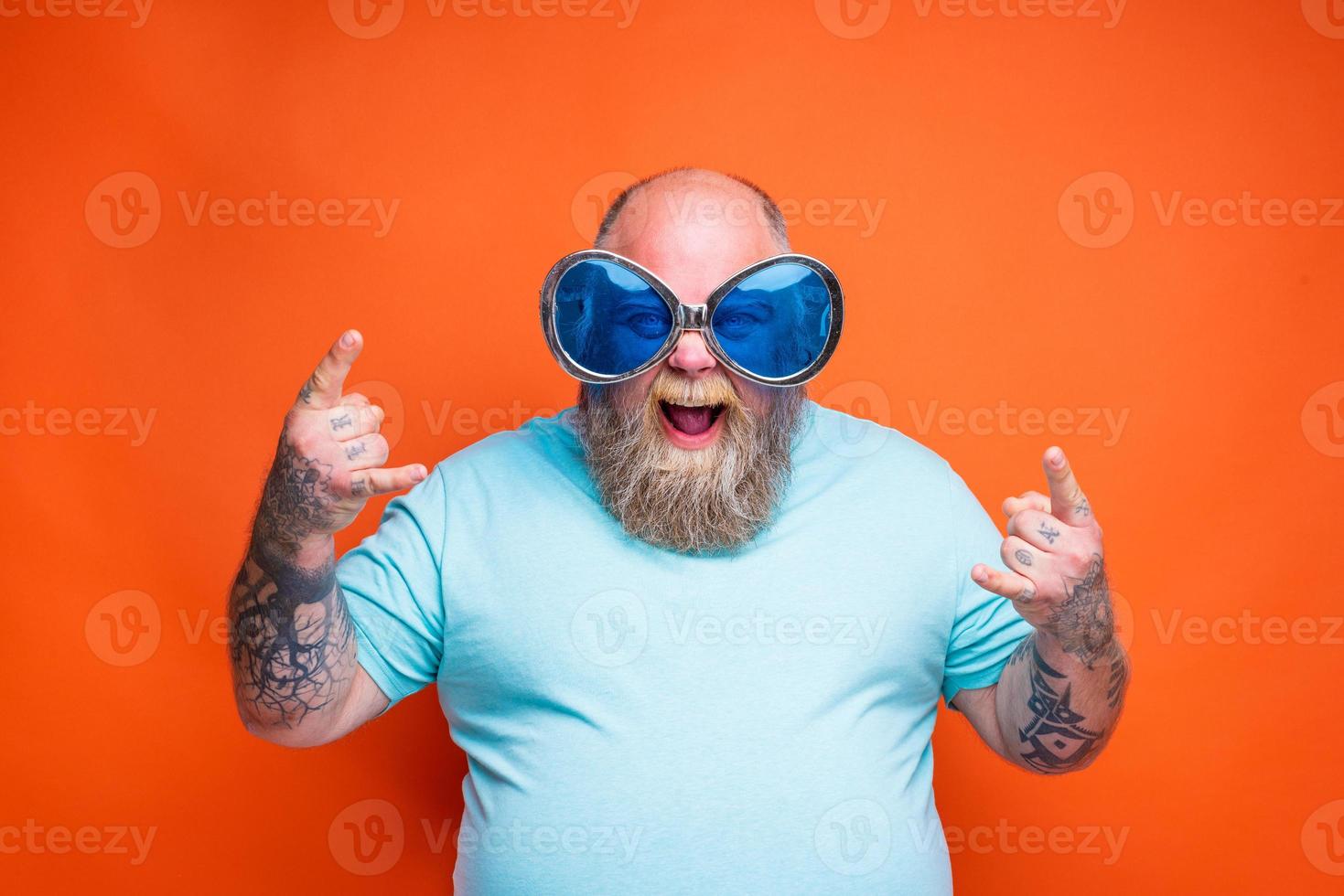 Grasso contento uomo con barba, tatuaggi e occhiali da sole fa il gesto di il corna foto