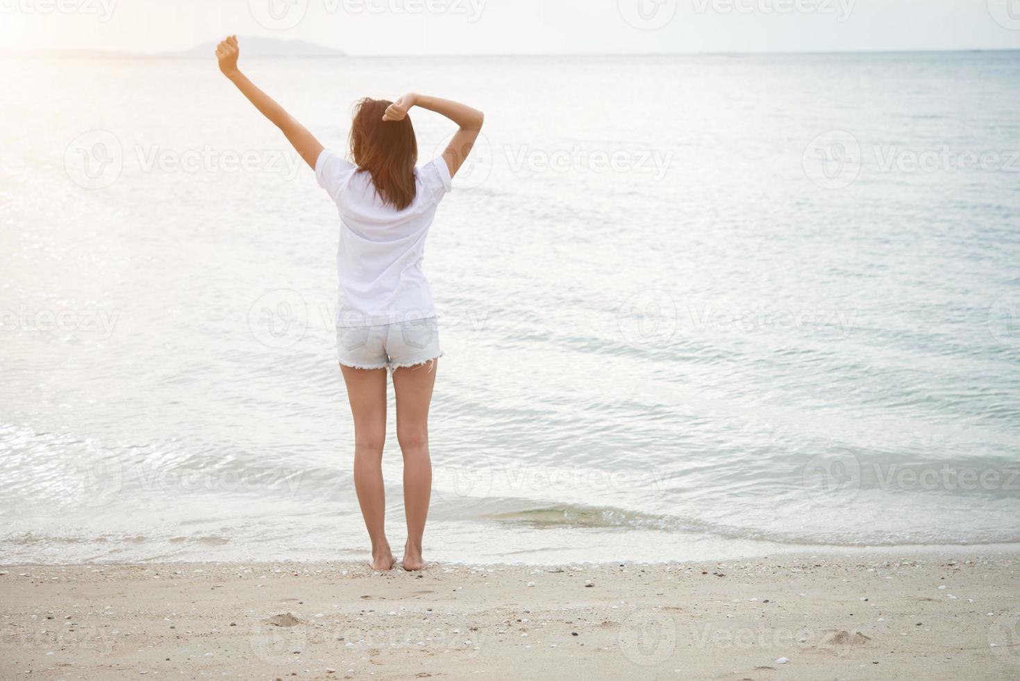 posteriore della giovane donna in piedi allungare le braccia in aria sulla spiaggia a piedi nudi foto