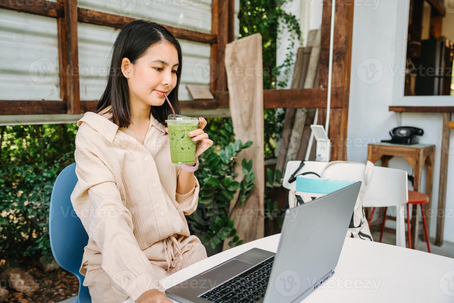 giovane asiatico donna potabile ghiacciato verde tè e Lavorando con il computer portatile a scrivania nel Giardino dietro la casa a caffè negozio foto