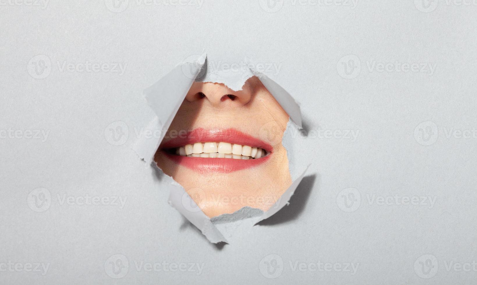 donna mostrando sua bocca con bianca trentadue denti scherzosamente nel strappato carta buco, attraverso sfondamento di grigio sfondo. cosmetici pubblicità concetto. oh, grande notizia foto