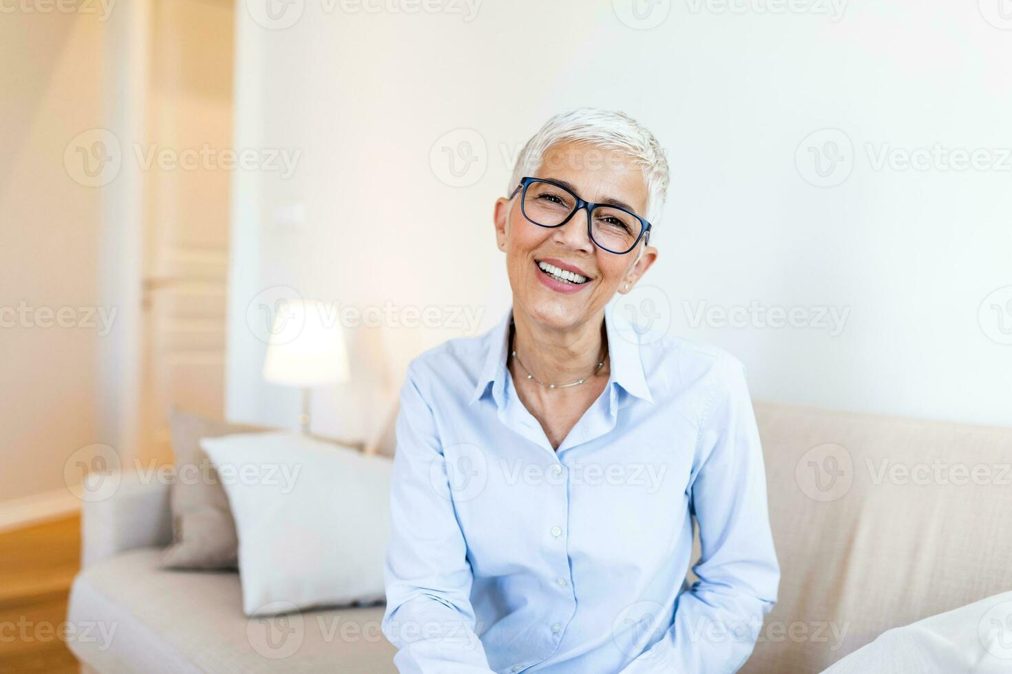 anziano donna con bicchieri sorridente mentre seduta su un' divano a casa. maturo donna sensazione contento foto