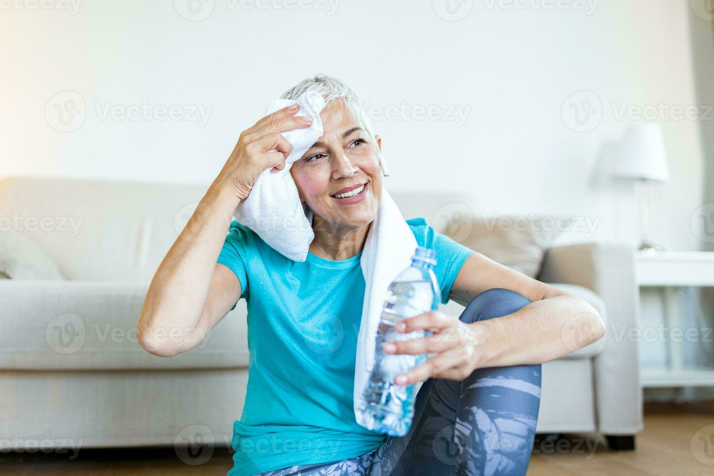 anziano donna Tenere plastica bottiglia di acqua, asciugandosi sudore con un' asciugamano, esausto dopo il quotidiano addestramento. anziano donna assunzione un' rompere mentre esercizio a casa. seduta su fitness stuoia e riposo foto