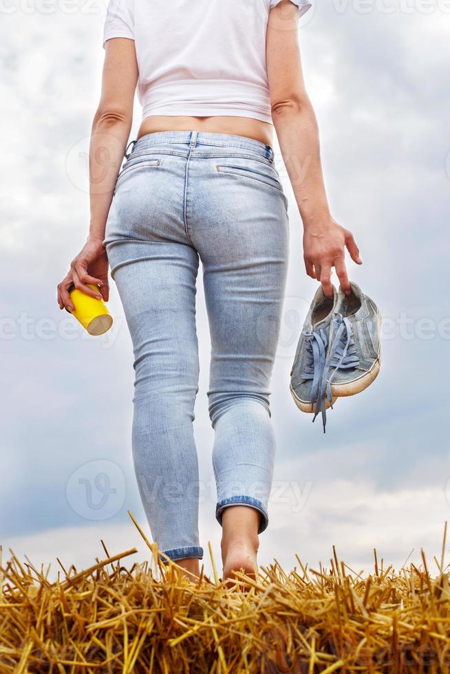 a piedi nudi ragazza con scarpe da ginnastica e cartone tazza con caffè nel mano In piedi nel il agricolo campo foto