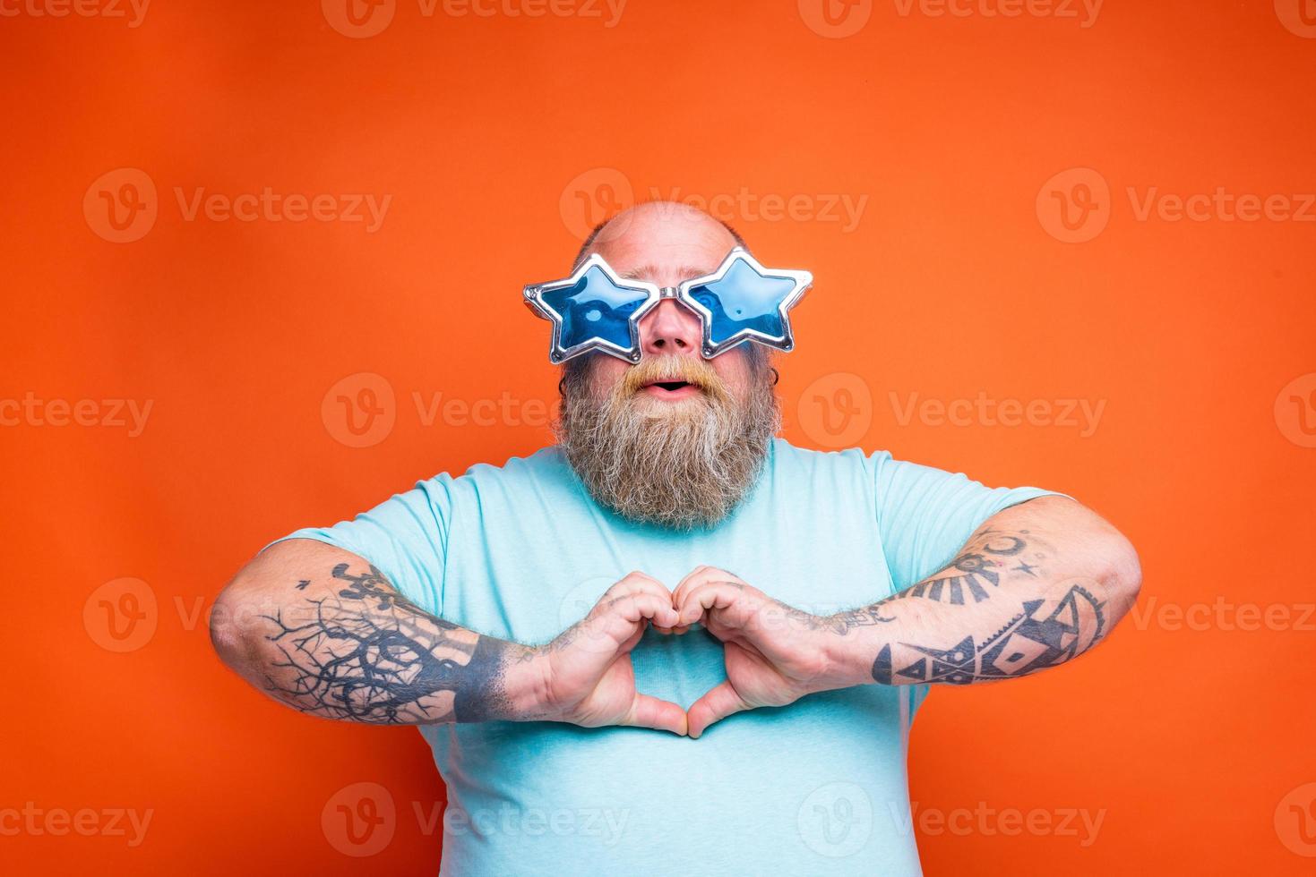 Grasso uomo con barba, tatuaggi e occhiali da sole fa cuore forma con mani foto