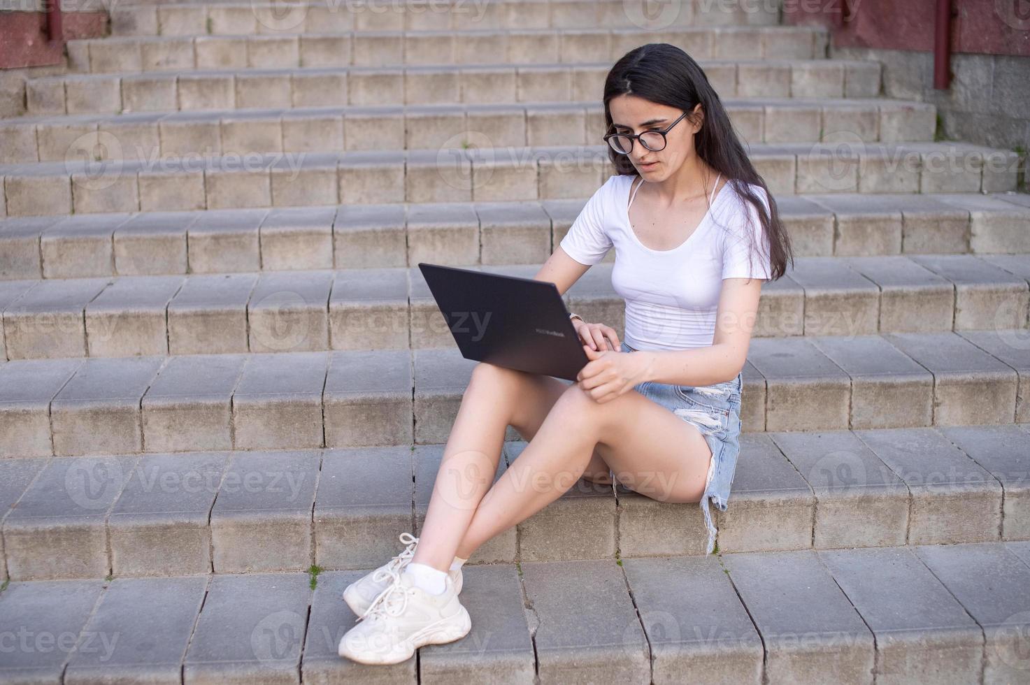 un' ragazza con bicchieri è seduta su il le scale con un' il computer portatile su sua giro foto