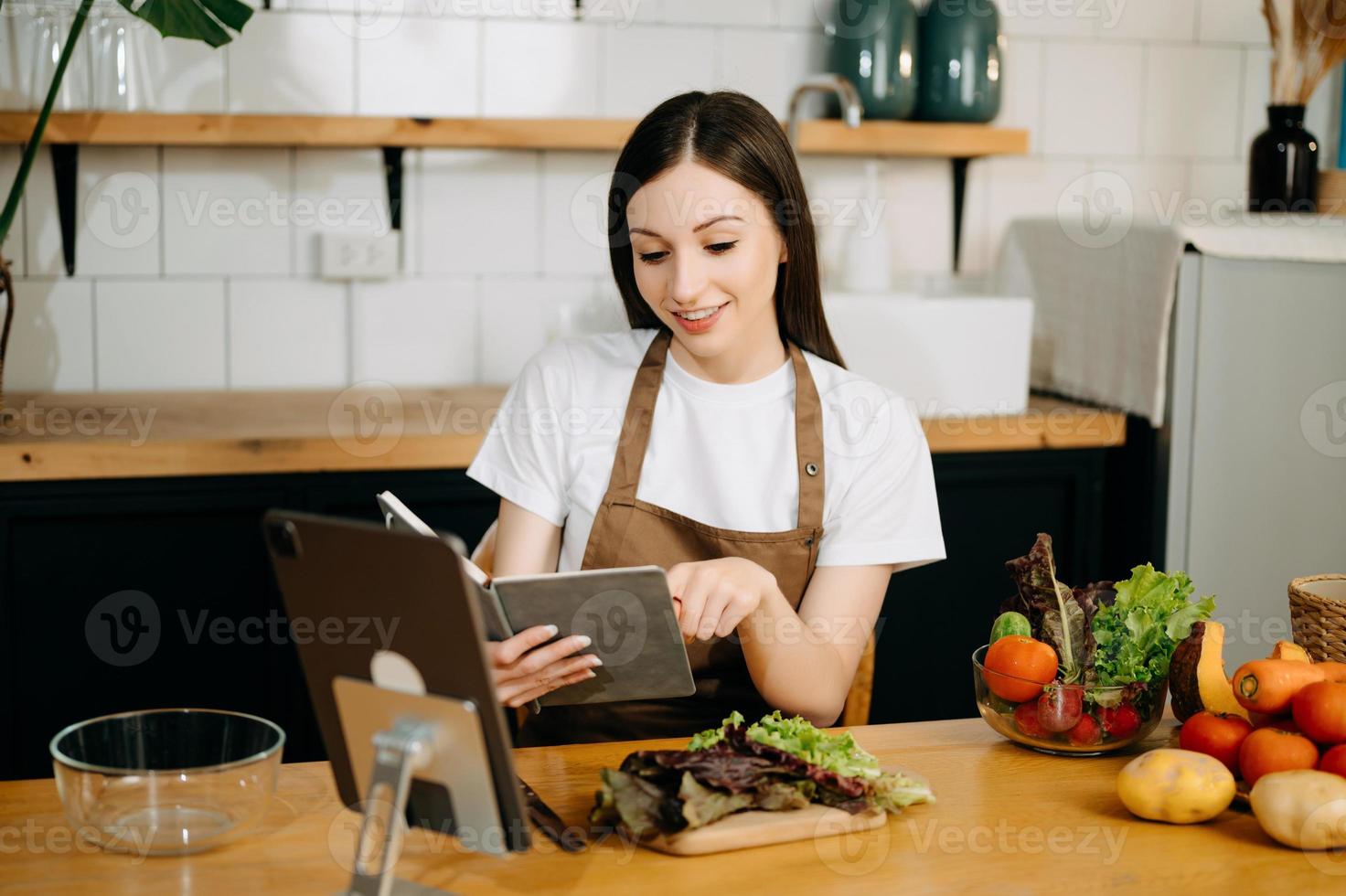 giovane bellissimo donna nel il cucina nel un grembiule, fresco verdure su il tavolo, scrive giù sua preferito ricette, arriva su con idee per piatti foto