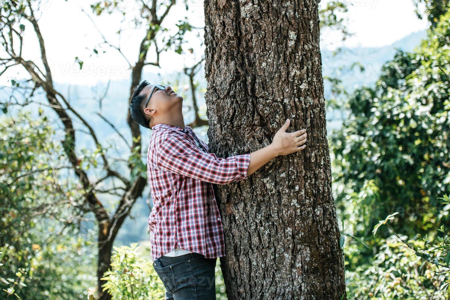 ritratto di contento asiatico uomo abbracciare un' albero nel foresta foto