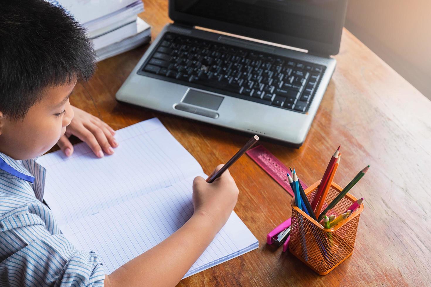 ragazzo che fa i compiti con notebook, laptop, cucitrice e una tazza di matite su una scrivania in legno foto