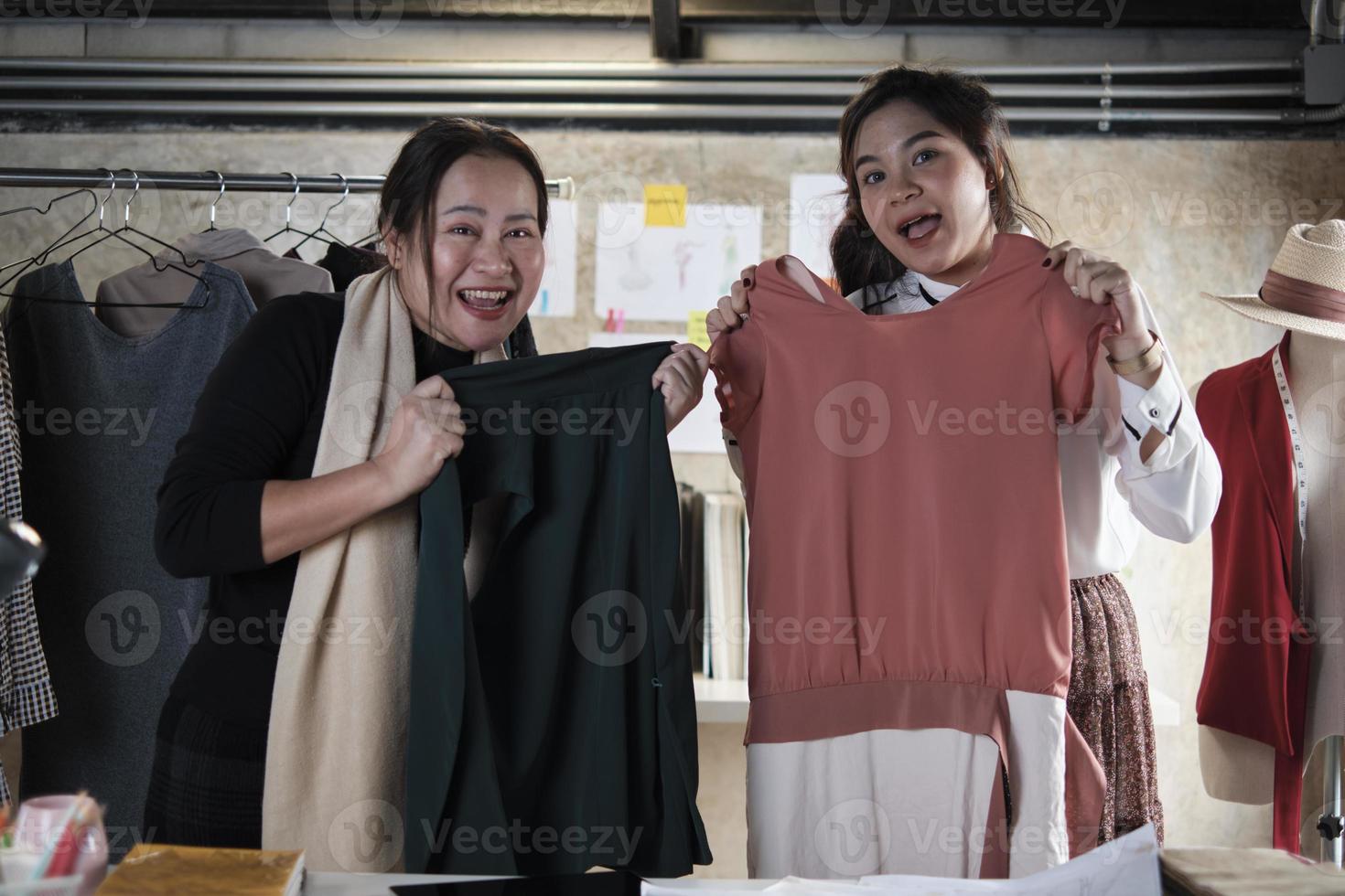 asiatico di mezza età femmina moda progettista e giovane adolescente apprendista sarto orgoglioso e allegro con indumenti design opera successo, mostrando vestiti lavoro nel studio, contento costume boutique piccolo attività commerciale. foto