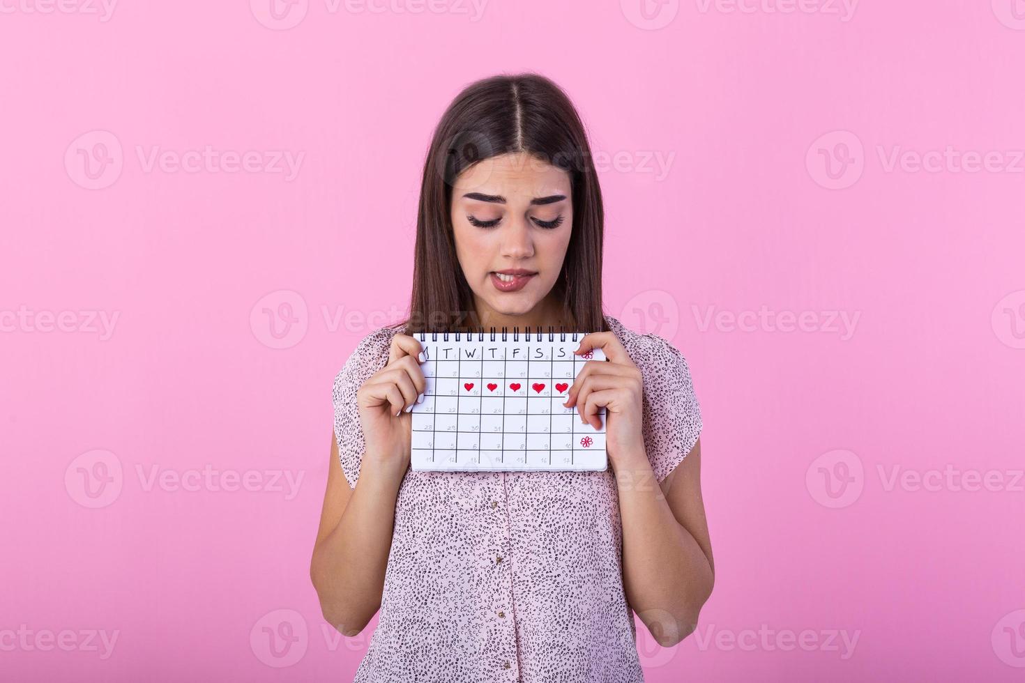 ritratto di imbarazzato carino ragazza pungente sua labbro e Tenere mestruale calendario con disegnato cuori per periodo e guardare a telecamera isolato al di sopra di rosa sfondo foto