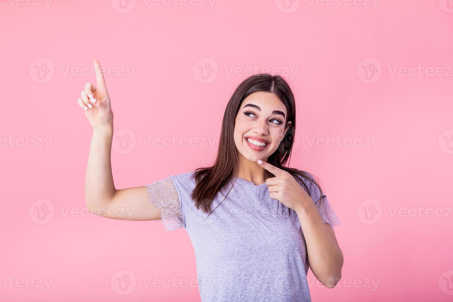 ritratto di brunetta donna con lungo capelli nel di base maglietta gioia e puntamento dito a copyspace isolato al di sopra di rosa sfondo foto