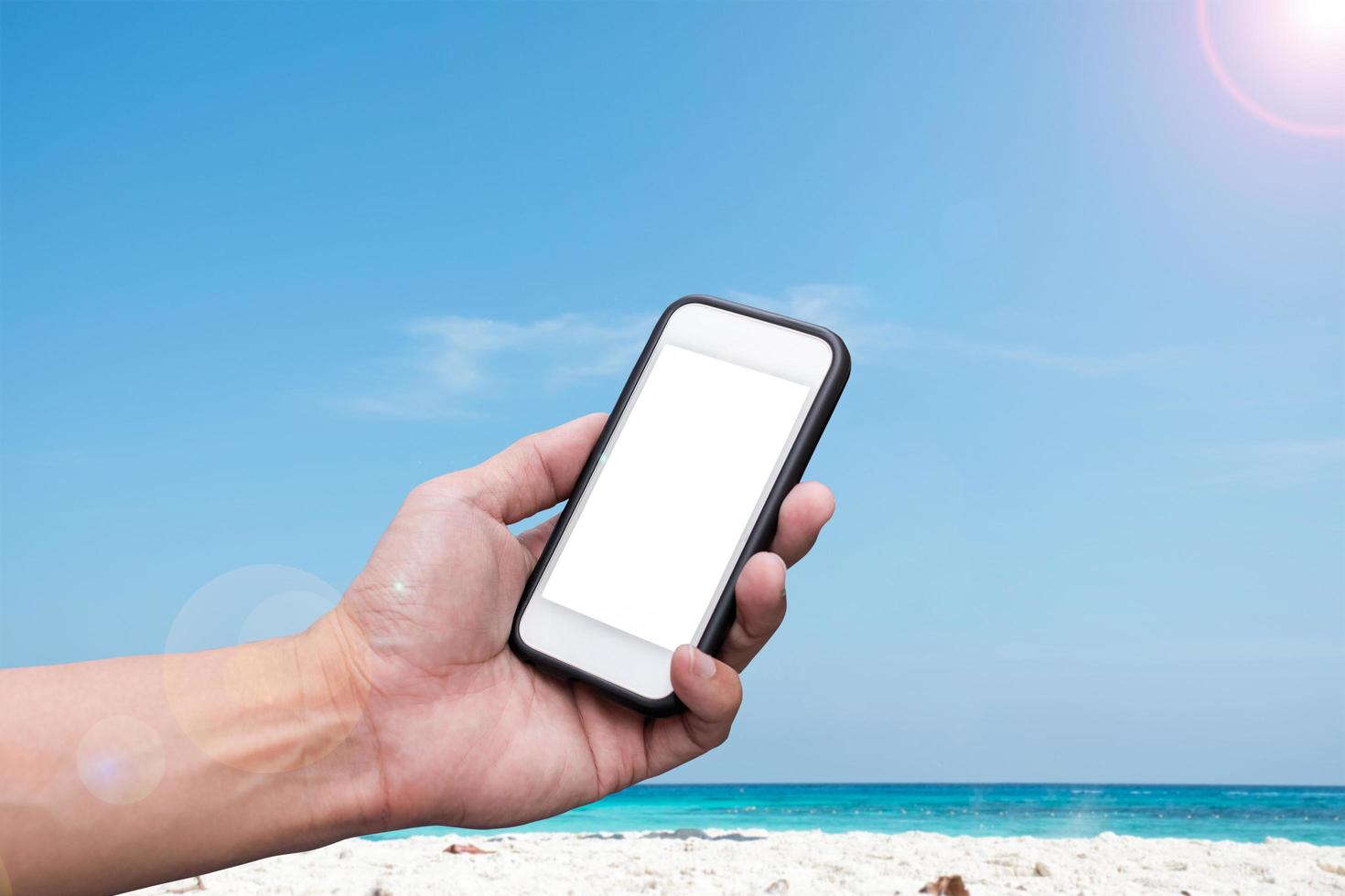 mano che tiene uno smartphone su una spiaggia e uno sfondo di cielo blu foto