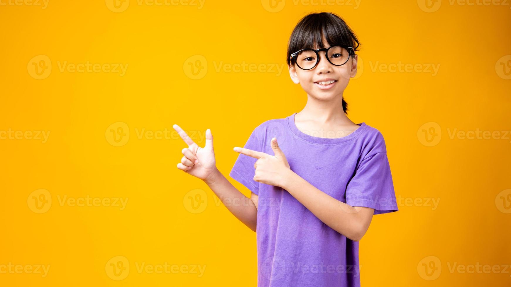 giovane ragazza asiatica in camicia viola e occhiali che punta a copia spazio in studio con sfondo giallo foto