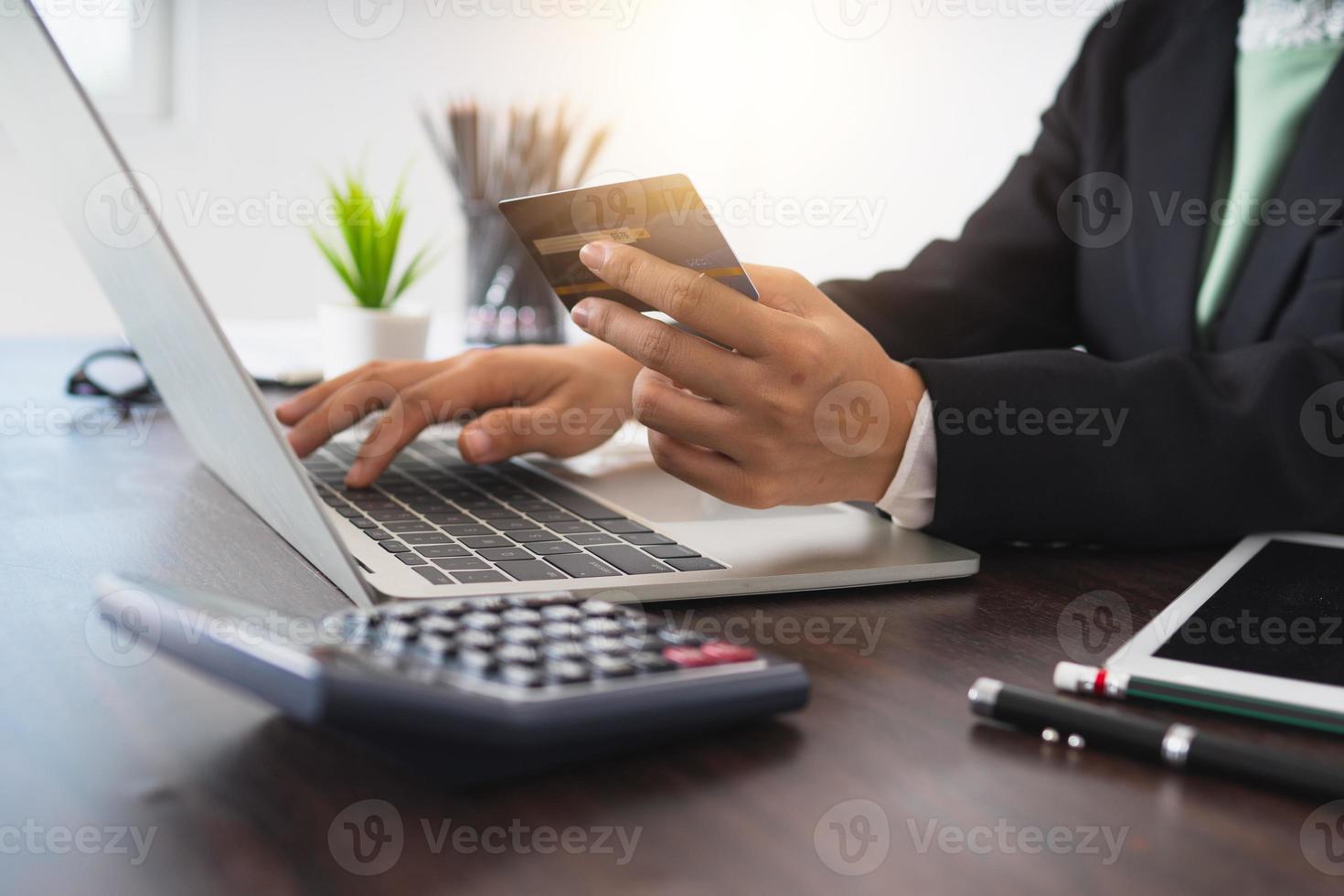 imprenditrice tenendo la carta di credito e digitando su un computer portatile accanto alla calcolatrice a una scrivania in legno marrone foto