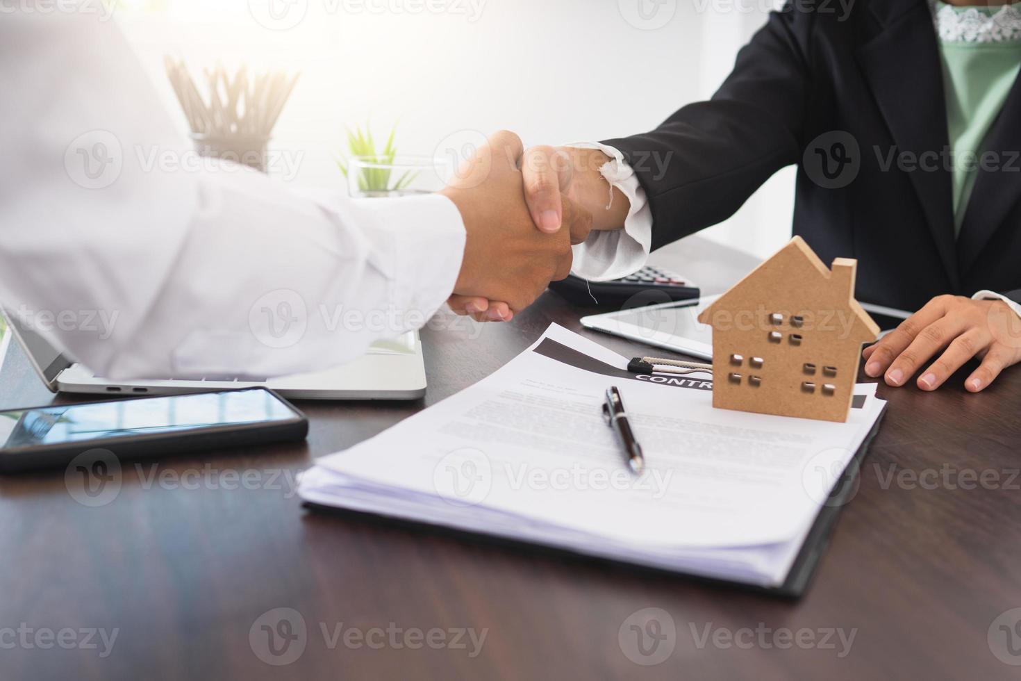 due persone si stringono la mano accanto a un modello di casa e un contratto foto