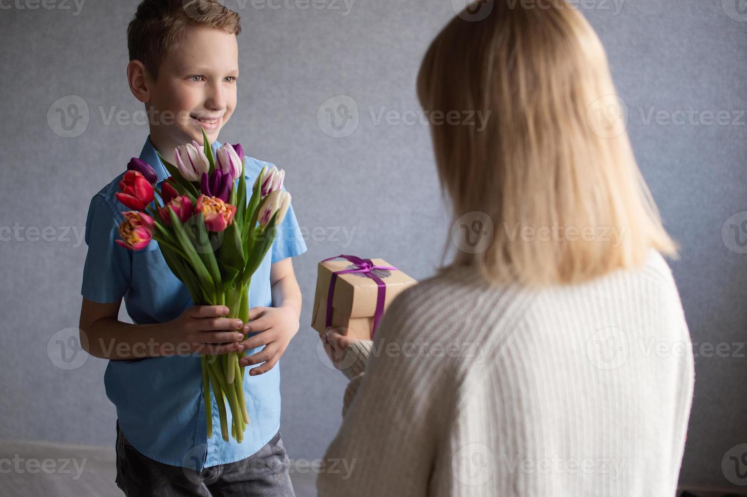 carino ragazzo nel un' blu camicia dà un' mazzo di tulipani e un' scatola con un' regalo per mamma foto