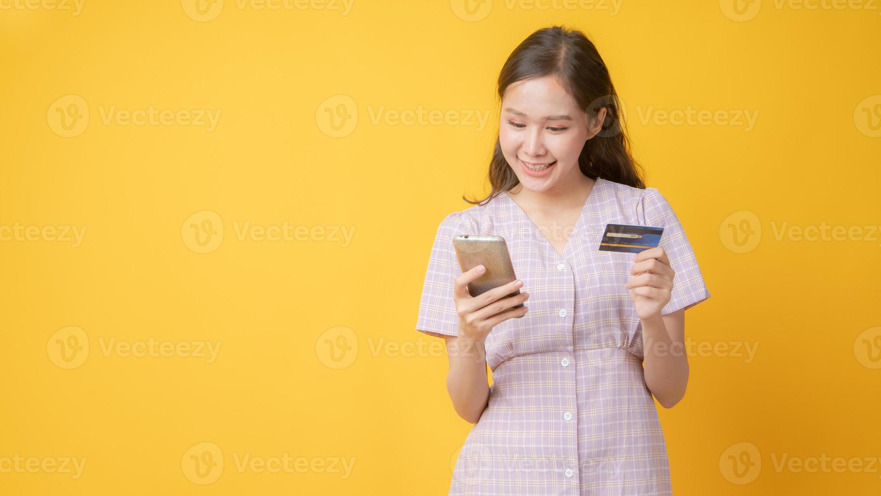 donna asiatica sorridente, tenendo la carta di credito e guardando il cellulare su sfondo giallo foto