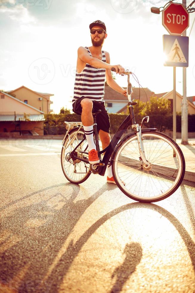 giovane uomo nel occhiali da sole equitazione un' bicicletta su un' città strada a tramonto leggero foto