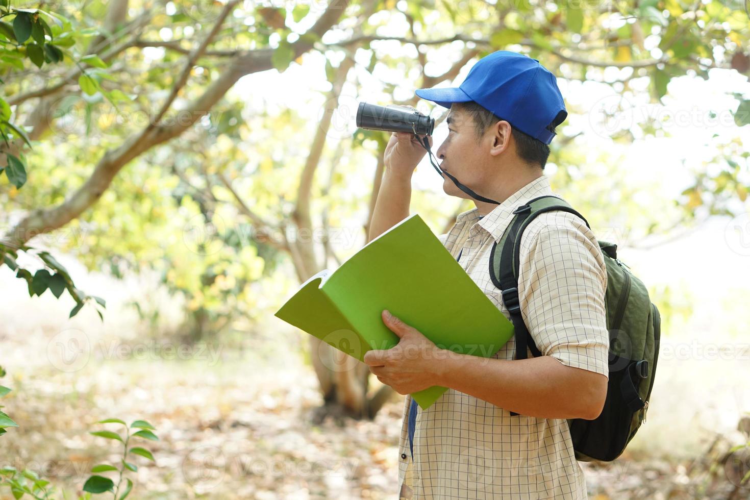 asiatico uomo esploratore indossa blu berretto, detiene binoculare nel foresta per sondaggio botanico impianti e creature animali selvatici. concetto, natura esplorazione. ecologia e ambiente. foto