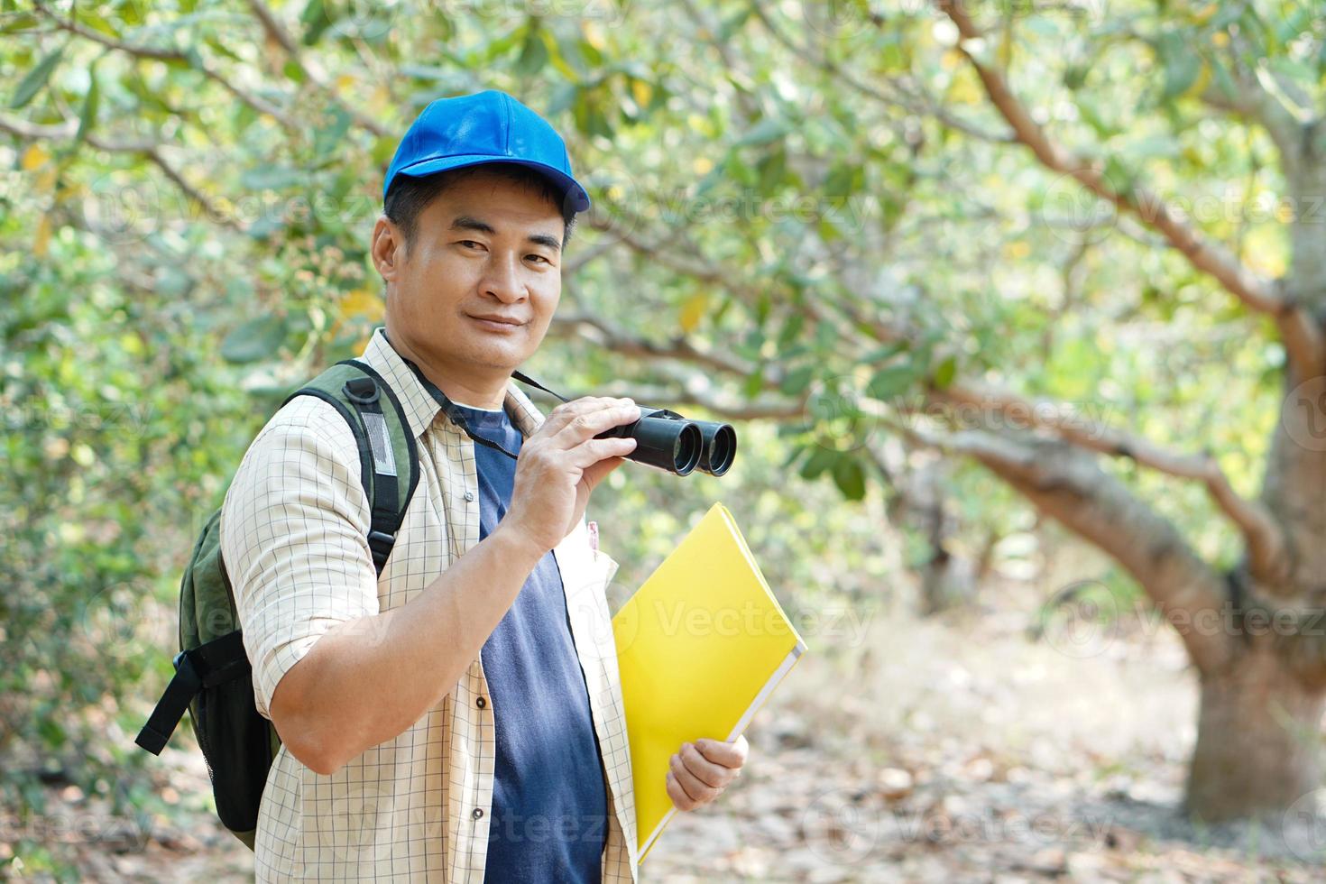 asiatico uomo esploratore indossa blu berretto, detiene binoculare nel foresta per sondaggio botanico impianti e creature animali selvatici. concetto, natura esplorazione. ecologia e ambiente. foto