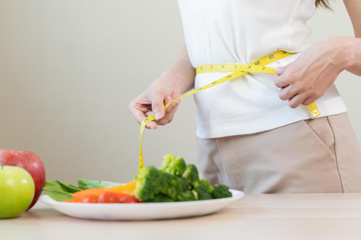 sottile forma asiatico giovane donna, ragazza in piedi, mano nel misurare nastro in giro vita, corpo sua avere un' verdure su tavolo come bianca sfondo. dieta sessione per benessere Salute, mangiare bene cibo le persone. foto