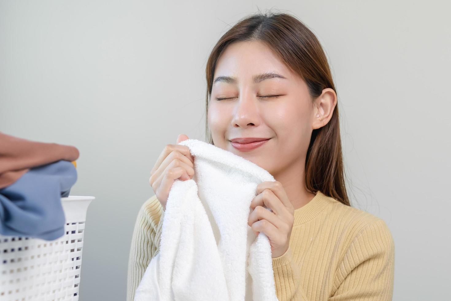 sentire morbidezza, sorridente asiatico giovane donna, ragazza toccante soffice asciugamano cotone, odorare fresco pulito Abiti su tavolo dopo lavaggio, lavanderia, asciutto. domestico Lavorando a casa. lavanderia e domestica. foto