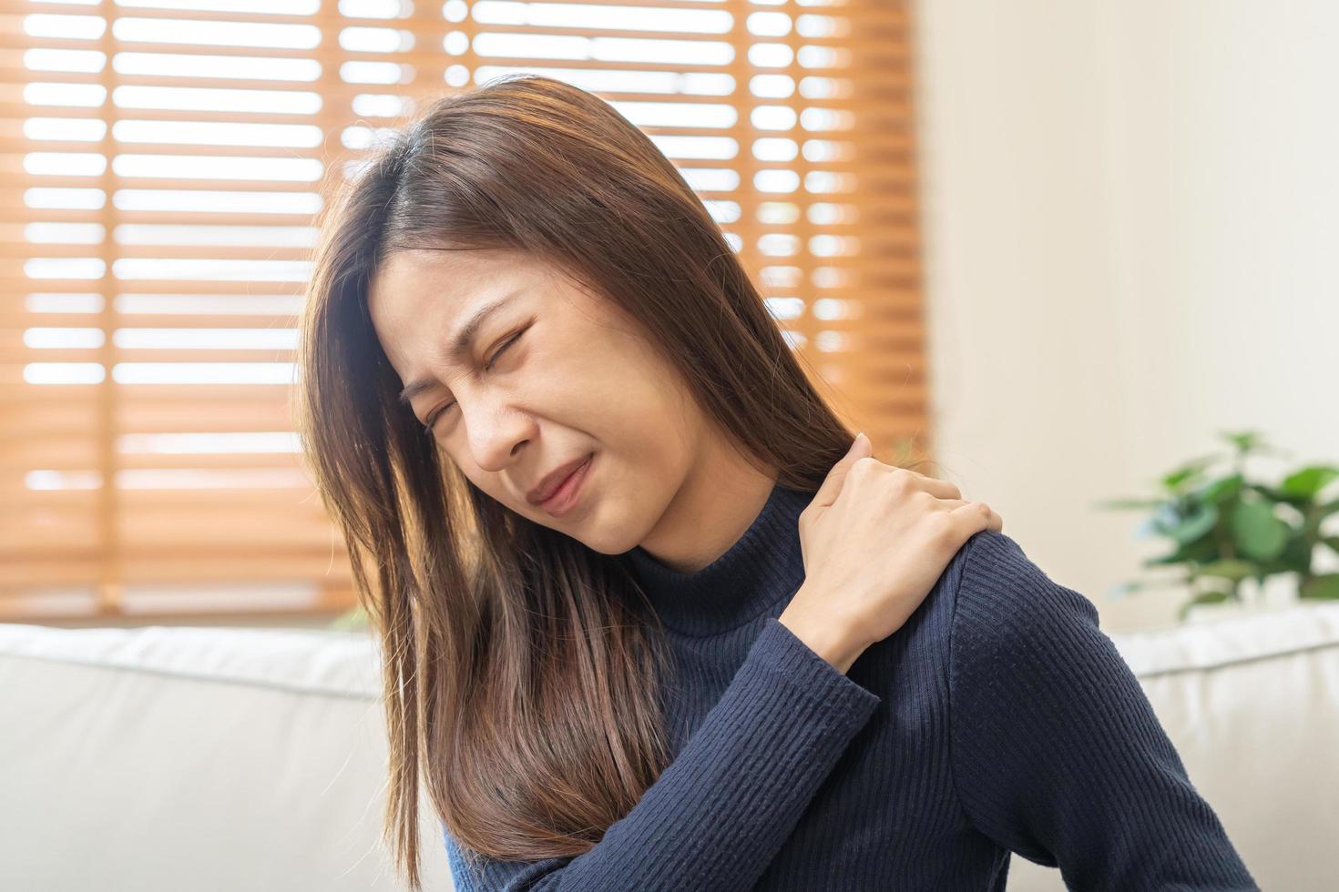 corpo muscoli rigido problema, asiatico giovane attraente donna, ragazza dolore con indietro dolore dolore a partire dal opera, Tenere massaggio sfregamento spalla male o irritato, doloroso seduta su divano a casa. assistenza sanitaria le persone. foto