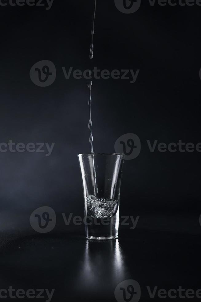 un' ruscello di alcool versa in un' bicchiere contro un' nero sfondo. acqua spruzzata. vicino su foto