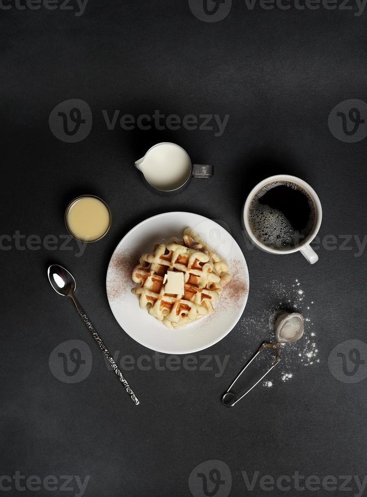 superiore Visualizza di gustoso cialde piatto, caramello salsa, caffè tazza, latte, cucchiaio da dessert, filtro su un' nero calcestruzzo sfondo foto