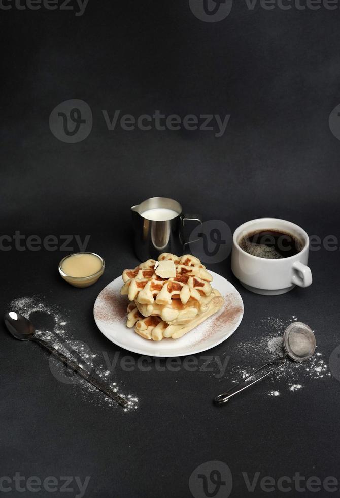 fatti in casa di prima colazione gustoso cialde, caramello salsa, caffè tazza, latte, cucchiaio da dessert, filtro su un' nero calcestruzzo sfondo foto