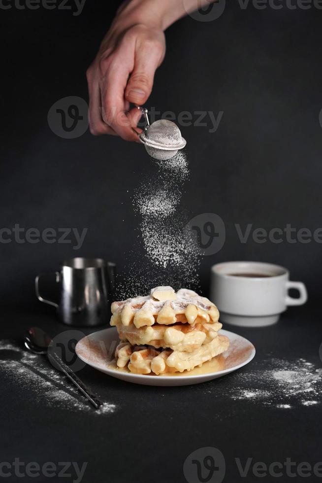 caucasico dell'uomo mano spruzzatori zucchero polvere su delizioso cialde, caramello salsa, caffè tazza, latte, dolce cucchiaio, filtro su nero calcestruzzo sfondo foto